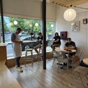 4 people in a coffee shop, 3 sitting and 1 wearing an apron, look off camera