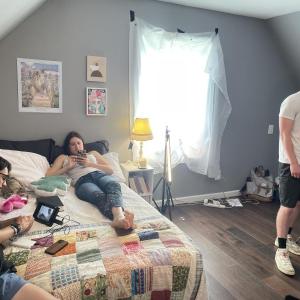 a standing man operates a camera while another one looks at a camera monitor and a woman lays nearby looking at her cell phone. 
