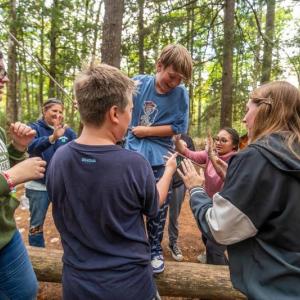 One student tries to balance as they walk across the log without holding onto a rope, although one is made available when needed, while their classmates stand ready to catch them or steady them if they lose their balance during a trust exercise at the 4-H Camp and Learning Center at Tanglewood in Lincolnville. )Photo courtesy of University of Maine Cooperative Extension)
c