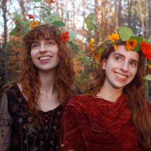 Two women with floral crowns. One in black dress, one in red dress.