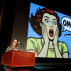 Big-screen comic scene; two women at lectern