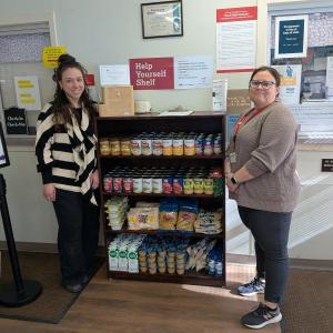 Two women stand next to shelving unit stocked with food