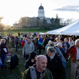 people gather on a lawn with the state house visible in the background