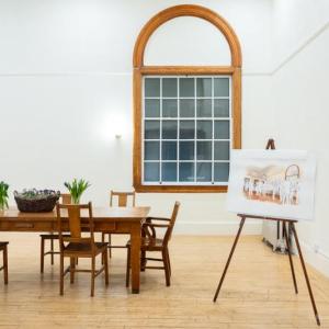 The boardroom at Watts Hall as viewed from the entrance. The room is brightly lit, with white walls, a wood floor, and a large wooden table with vases of fresh cut tulips. There is a large window with an arched top; it's dark outside. There is an easel in the corner, displaying an artist's rendering of what the room might look like filled with people mingling during an event. Photo credit: Nicole Langille Imagery.