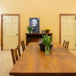 This image shows the Watts Hall boardroom as viewed from inside. In the foreground is a large wooden table with vases of fresh cut tulips. The back wall is a rich, golden color, and there are two doors in the wall, one leading to a closet, the other to the prep kitchen. There's a black and white photo of Captain Samuel Watts propped up on a credenza against the wall, and it's like he's looking out over the room. Photo credit: Nicole Langille Imagery.