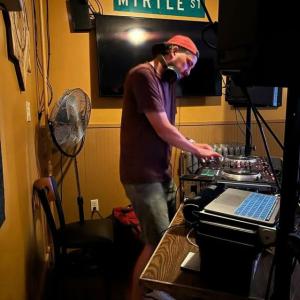 A picture of a man, DJ Souz, standing in front of DJ equipment. Hanging on the wall above his head is a road sign that says Myrtle St.
