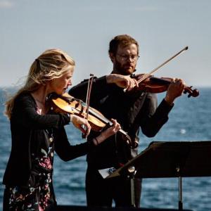 woman, man play string instruments against ocean backdrop
